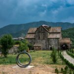 Explore an ancient church set against lush mountains with an adjacent cemetery and a striking sculpture.